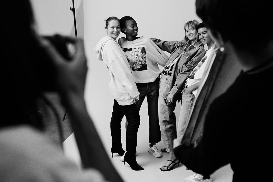 Black-and-white behind-the-scenes shot from an H&M campaign: four models laughing together on a white studio set, framed by crew in the foreground